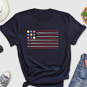May include: A navy blue t-shirt with a graphic of a pool table with balls and cues arranged in the shape of an American flag.