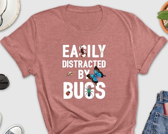 May include: A heather pink t-shirt with the text "EASILY DISTRACTED BY BUGS" in white. The text is surrounded by images of a ladybug, a butterfly, a spider, and a fly.