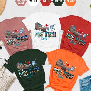 May include: A collection of short-sleeved t-shirts in various colors, including heather mauve, white, red, forest green, and orange. Each shirt features a graphic design with the text "MRI TECH Crew" and an illustration of medical equipment.