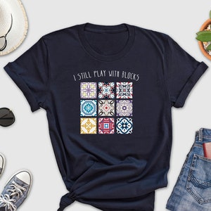 May include: Navy blue t-shirt with a white graphic that says "I still play with blocks". The graphic features a colorful pattern of nine square tiles.