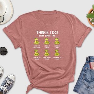 May include: A pink t-shirt with the text "THINGS I DO IN MY SPARE TIME" and a series of illustrations of snakes with text describing activities related to snakes.