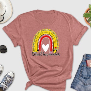 May include: A heather pink t-shirt with a rainbow made of yellow pencils with red erasers and the alphabet printed on them. A white heart is in the center of the rainbow. The text "School bus monitor" is printed below the rainbow.