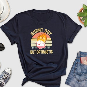 May include: A navy blue t-shirt with a vintage-style graphic of a burnt marshmallow with the text "Burnt Out But Optimistic".