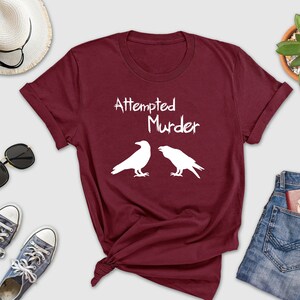 May include: Burgundy t-shirt with white text that reads "Attempted Murder" and two white silhouettes of ravens.