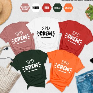 May include: A collection of short-sleeved t-shirts in various colors, including white, red, orange, and forest green. Each shirt features the text "SPD CREW" in a white, stylized font. The shirts are displayed with accessories like sunglasses and a straw hat.