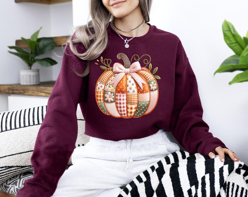 A woman wearing a burgundy sweater with a patchwork pumpkin design sits on a striped pillow.