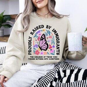 A woman wearing a beige sweatshirt with a graphic design and text printed on it, sitting on a couch with a mug in her hand.