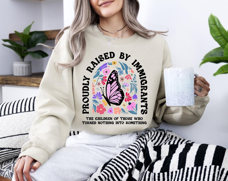 A woman wearing a beige sweatshirt with a graphic design and text printed on it, sitting on a couch with a mug in her hand.