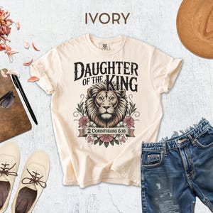 a casual outfit consisting of a white t-shirt with a lion graphic, denim shorts, and a brown hat, along with some accessories such as sunglasses, a notebook, and a pair of shoes.