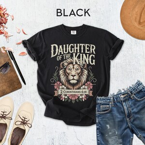 a casual outfit consisting of a black t-shirt with a lion graphic, distressed denim shorts, a brown hat, sunglasses, a notebook, and a pair of white sneakers.