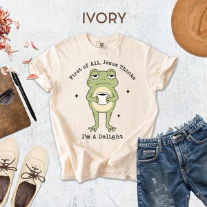 a white t-shirt with a cartoon frog holding a cup, a pair of blue jeans, a brown hat, sunglasses, a notebook, and a pair of white shoes.