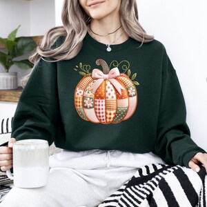 A woman wearing a green sweater with a patchwork pumpkin design is sitting on a striped pillow, holding a white mug.