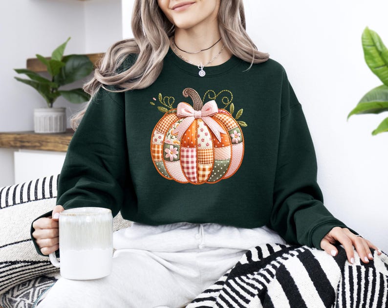 A woman wearing a green sweater with a patchwork pumpkin design is sitting on a striped pillow, holding a white mug.