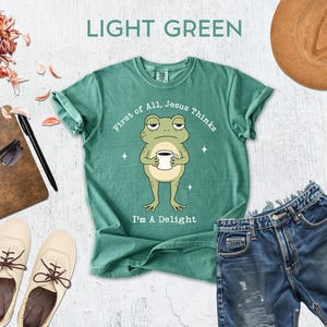 a green t-shirt with a cartoon frog, a pair of denim shorts, a hat, sunglasses, a notebook, and a cup, all arranged on a white background.