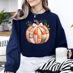 A woman wearing a navy blue sweater with a patchwork pumpkin design sits on a striped blanket, holding a white mug.