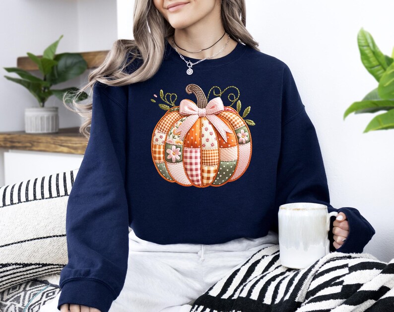 A woman wearing a navy blue sweater with a patchwork pumpkin design sits on a striped blanket, holding a white mug.