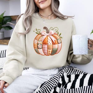 A woman wearing a beige sweater with a pumpkin embroidered on it, holding a mug, and sitting on a striped blanket.