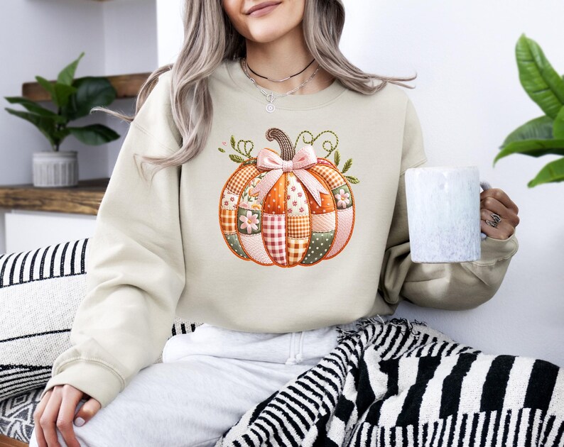 A woman wearing a beige sweater with a pumpkin embroidered on it, holding a mug, and sitting on a striped blanket.