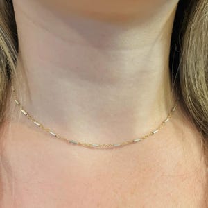 May include: A delicate gold chain necklace featuring evenly spaced, small, cylindrical silver-toned metal beads. The necklace rests closely around the neck, showcasing a minimalist design with alternating gold chain links and smooth silver-toned accents.