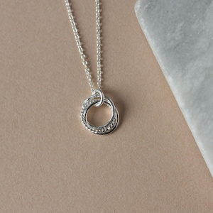 May include: A silver necklace with a pendant featuring three interlocking rings. The rings are textured and have a polished finish.