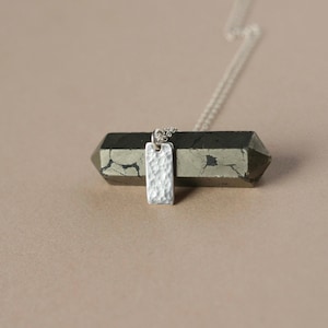 May include: A silver pendant necklace with a textured rectangular charm. The charm hangs from a delicate chain. The necklace is displayed on a dark, geometric stone against a neutral background. The pendant has a hammered metal appearance.