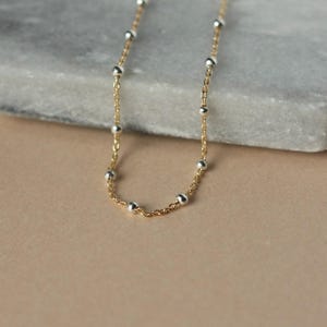 May include: A delicate gold necklace featuring small silver beads evenly spaced along the chain. The necklace is displayed on a neutral surface, highlighting its simple elegance. The chain is approximately 40 cm long.