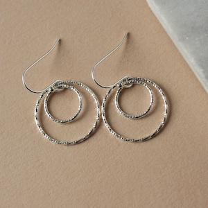May include: A pair of silver hoop earrings. Each earring has two textured, concentric circles suspended from a hook. The earrings are displayed on a neutral-toned surface.