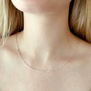 May include: A silver chain necklace with a delicate, linked design. The chain is worn by a person with long blonde hair.