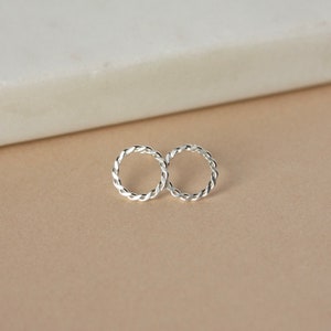 May include: A pair of silver-coloured, braided hoop earrings. The earrings are circular with a woven design, and are displayed on a neutral background. The earrings are a simple, elegant accessory.