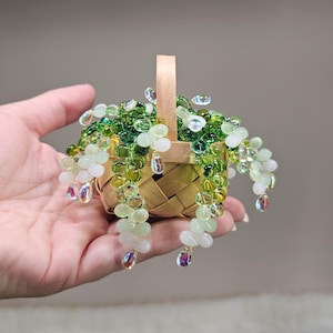 Peut inclure: Un panier miniature en bois tressé contenant une plante décorative composée de perles de verre translucides dans des tons vert clair, vert foncé et blanc. Les vignes perlées retombent sur les côtés du panier avec des accents en goutte d'eau irisés.
