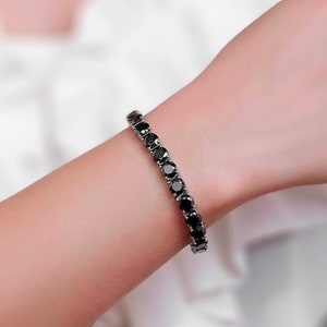 May include: A black diamond tennis bracelet with a silver chain. The bracelet is on a person's wrist.