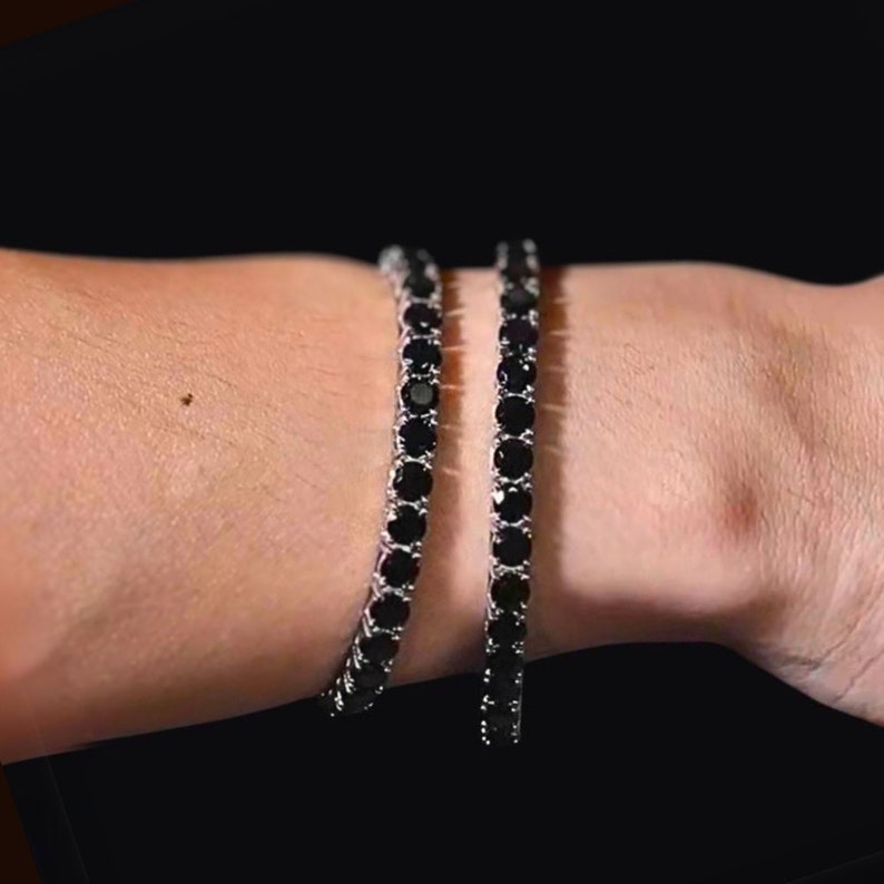 May include: Two black diamond tennis bracelets on a person's wrist. The bracelets are made of silver and have a row of black diamonds.