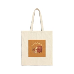 May include: Cream-colored tote bag with a square graphic. The graphic features a cozy illustration of a red armchair, a side table with a coffee cup, and a blanket. The text "Home is My Happy Place" is written above the illustration.