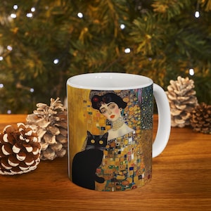 May include: White ceramic mug with a colorful image of a woman with a black cat. The image is a reproduction of a famous painting. The woman is wearing a gold dress and a pearl necklace. The cat is sitting on her lap. The background is a mosaic of gold, black, and white squares.