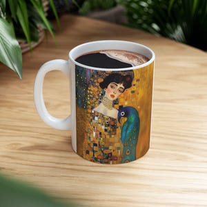 May include: White ceramic mug with a colorful image of a woman with a parrot on her shoulder. The image is a mosaic of gold, blue, green, and white squares. The mug is filled with coffee.