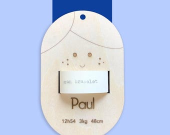 Puede incluir: Una placa ovalada de madera con una cara de dibujos animados, el texto "mon bracelet", el nombre "Paul" y las medidas "12h54 3kg 48cm".
