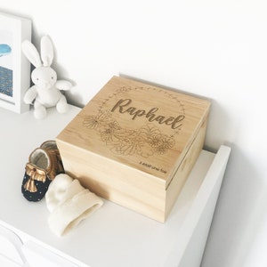Peut inclure: Un coffret en bois clair avec le nom "Raphaël" gravé sur le couvercle, entouré d'un motif floral. La boîte est rectangulaire avec un dessus légèrement incurvé. Un petit bonnet blanc et des chaussures de bébé sont à côté.