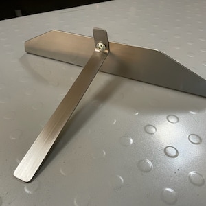 Pode incluir: Uma pá de pizza de metal prateado com uma alça longa. A pá tem uma lâmina plana e retangular com uma borda ligeiramente curva.