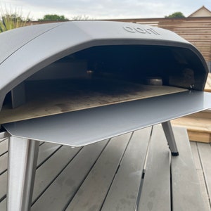 Puede incluir: Un horno de pizza gris para exteriores con frente y patas de acero inoxidable. El horno tiene una parte superior curva y una abertura rectangular. Se ve una piedra para pizza en el interior. El horno está sobre una terraza de madera.
