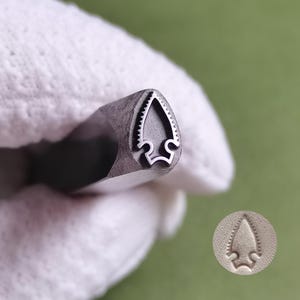 Op de afbeelding: Een metalen stempelgereedschap met een puntig pijlpuntontwerp met gekartelde randen. De stempelkop is gemaakt van donker staal en creëert een schone, verzonken afdruk van de pijlpuntvorm, die als voorbeeld op een licht oppervlak wordt getoond.