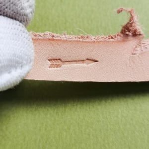 Op de afbeelding: Een strook lichtbruin, natuurkleurig leer met een klein, gestempeld pijlicoon. Het leer heeft een zichtbare, getextureerde nerf en ruwe, vezelige randen. Het pijlmotief is direct in het oppervlak van het materiaal gedrukt.