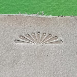 May include: A light beige piece of leather featuring a stamped sunburst pattern. The design consists of nine radiating teardrop-shaped impressions, each with a small circular indentation at the rounded end, creating a semi-circular fan motif.