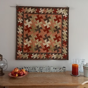 Peut inclure: Une courtepointe murale avec un motif géométrique en tissu brun, rouge et pêche. La courtepointe est accrochée à un mur blanc au-dessus d'une table en bois avec une lanterne, des fruits et des objets décoratifs.