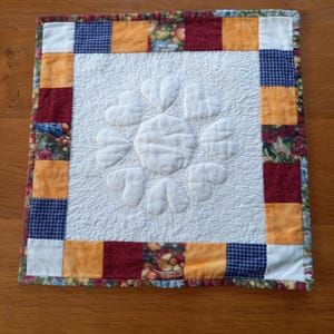 Peut inclure: Un set de table matelassé avec un centre blanc orné d'un motif en forme de cœur. La bordure est composée de carrés de différentes couleurs, dont l'orange, le rouge et le bleu, avec un bord à motifs floraux. Le set de table est posé sur une surface en bois.