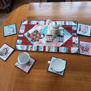 Peut inclure: Un service à thé matelassé comprenant une théière, des tasses et des dessous de verre. La théière est blanche avec un motif floral, et le set de table et les dessous de verre matelassés présentent un motif patchwork avec des motifs sur le thème du thé en rouge, bleu et rose.