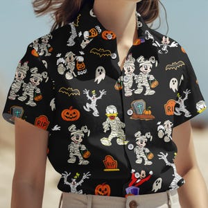 May include: Black button-up shirt with a Halloween-themed pattern. The design features cartoon characters dressed as mummies, pumpkins, bats, ghosts, and tombstones. The shirt has short sleeves and a collared neckline.