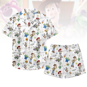 Könnte beinhalten: Weißes Pyjama-Set im Toy Story-Design. Das Set besteht aus einem kurzärmeligen Hemd und Shorts, die beide mit Figuren wie Woody und Buzz Lightyear sowie Sternen und Raketen bedruckt sind. Das Set ist bequem und hat einen klassischen Pyjama-Stil.