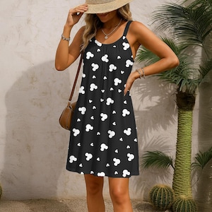 Op de afbeelding: Zwarte zomerjurk met een wit Mickey Mouse-hoofdpatroon. De jurk heeft een ronde hals, spaghettibandjes en een zoom tot op de knie. Het model draagt een strohoed en een bruine schoudertas.