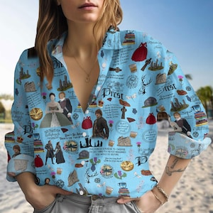 Puede incluir: Camisa abotonada azul claro con un collage de ilustraciones y texto. La camisa presenta imágenes de personas, castillos y otros símbolos, con texto que incluye "Prest" y "Lallybroch". La camisa tiene mangas largas y cuello.