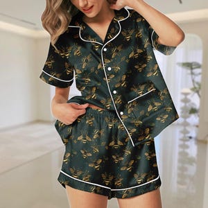 以下が含まれることがあります： ミツバチ柄のダークグリーンのパジャマセット。半袖トップスとショートパンツには、金色のミツバチと白いトリムがあしらわれています。ボタン留めのフロントとゆったりとしたフィット感で、快適な寝間着に最適です。
